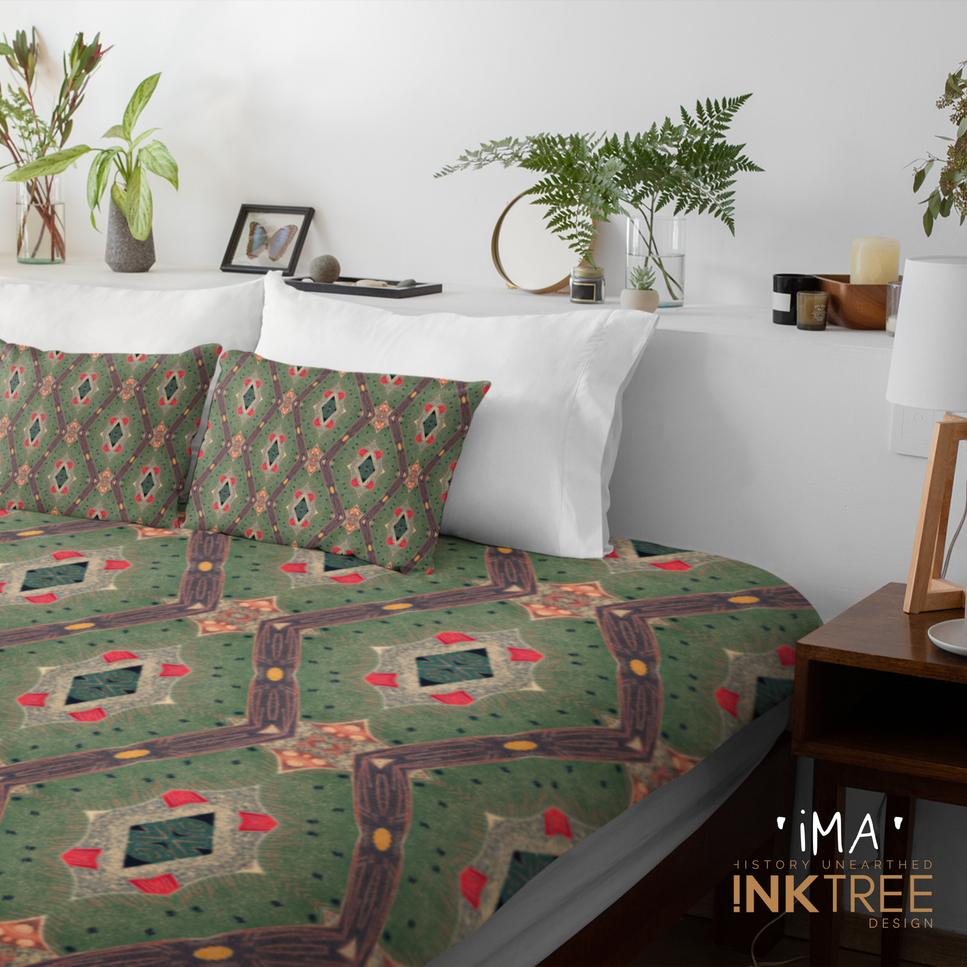 A quilt or doona cover and front pillows with a gold, black, red, green and white oriental looking pattern on a bed with a white wall, white pillows and white backboard background. There is a lamp with a white shade and wood base and plants and a small round metal mirror on the back board. There is also a ima, history unearthed ink tree design logo in the bottom right hand corner on it.
