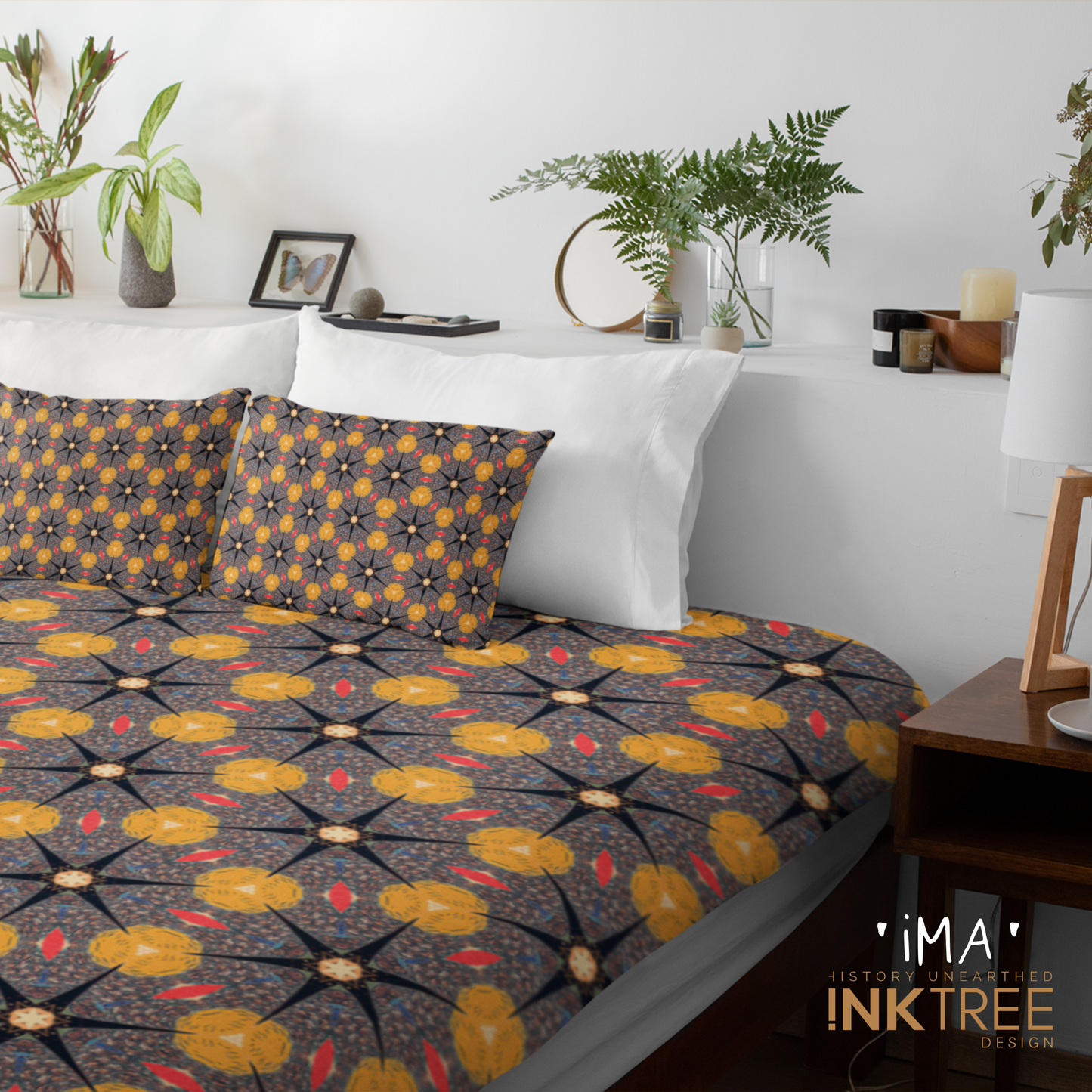 A quilt or doona cover and front pillows with a gold, black, blue, red and white oriental looking pattern on a bed with a white wall, white pillows and white backboard background. There is a lamp with a white shade and wood base and plants and a small round metal mirror on the back board. There is also a ima, history unearthed ink tree design logo in the bottom right hand corner on it.