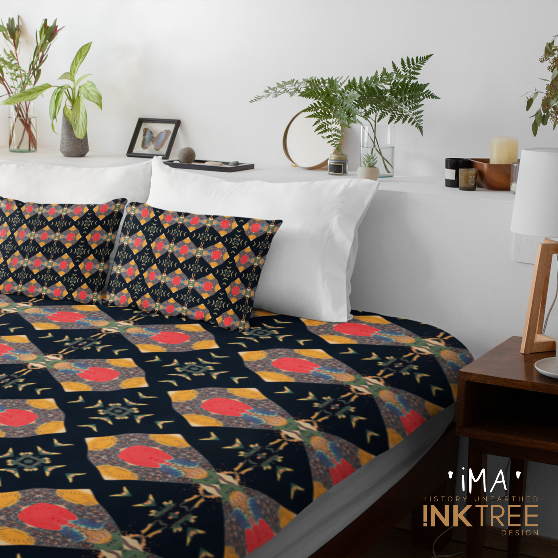 A quilt or doona cover and front pillows with a gold, black, blue, red, green and white oriental looking pattern on a bed with a white wall, white pillows and white backboard background. There is a lamp with a white shade and wood base and plants and a small round metal mirror on the back board. There is also a ima, history unearthed ink tree design logo in the bottom right hand corner on it.