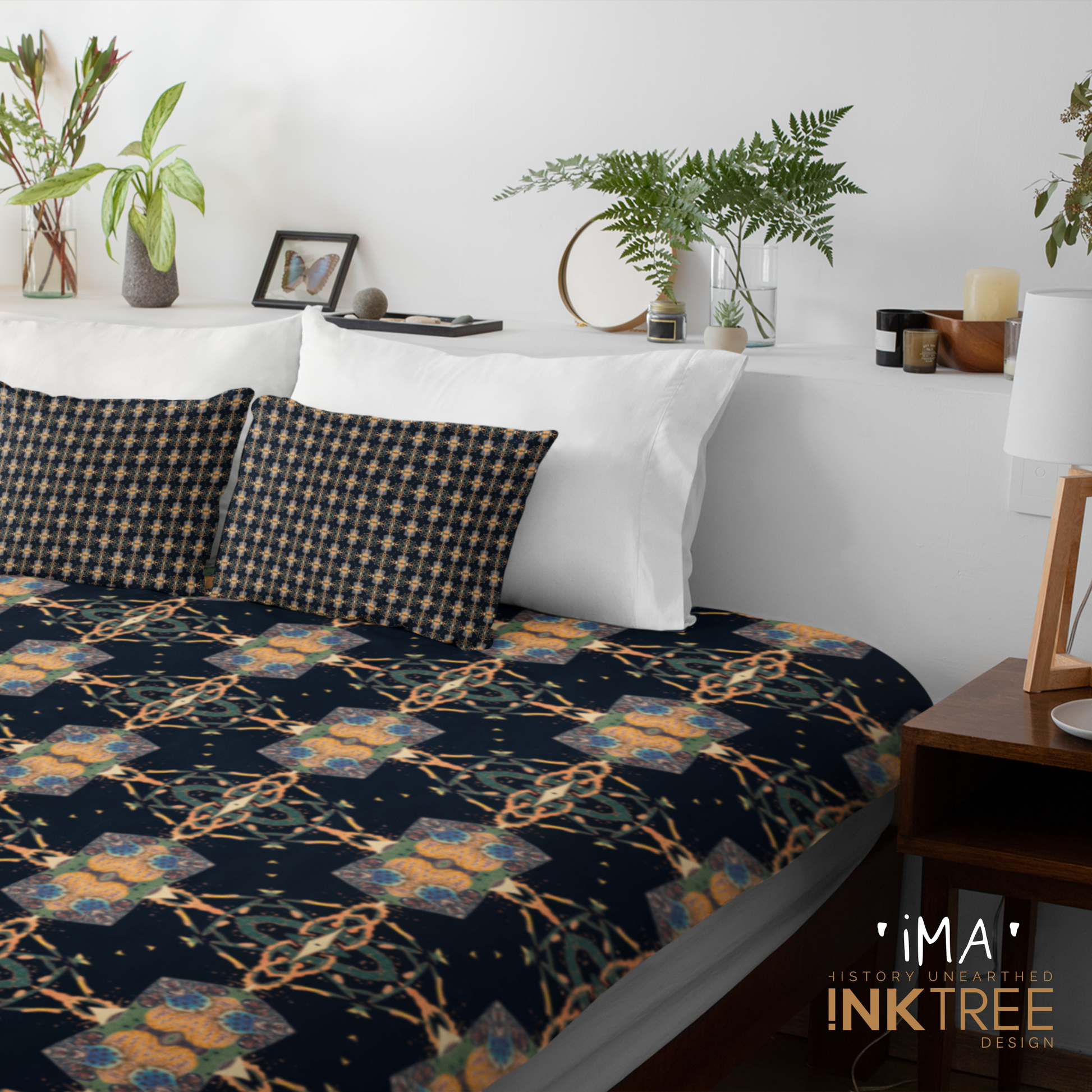 A quilt or doona cover and front pillows with a gold, black, blue, green and white oriental looking pattern on a bed with a white wall, white pillows and white backboard background. There is a lamp with a white shade and wood base and plants and a small round metal mirror on the back board. There is also a ima, history unearthed ink tree design logo in the bottom right hand corner on it.