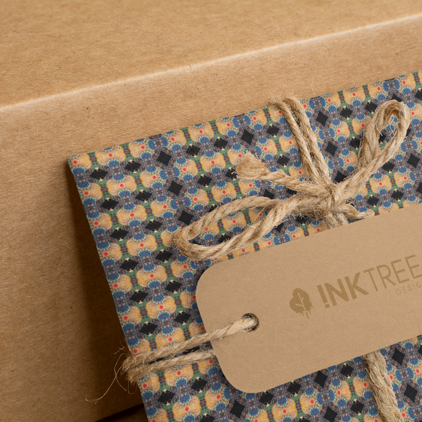 A present wrapped with a gold, black, blue, green red and white oriental looking pattern, tied with brown string with a brown paper tag with ink tree design logo on it, leaning on a brown paper background.