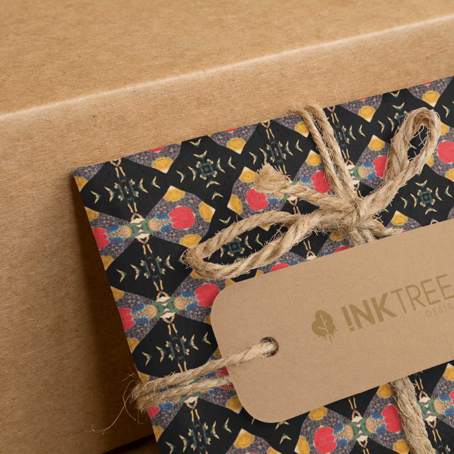 A present wrapped with a gold, black, blue, green red and white oriental looking pattern, tied with brown string with a brown paper tag with ink tree design logo on it, leaning on a brown paper background.