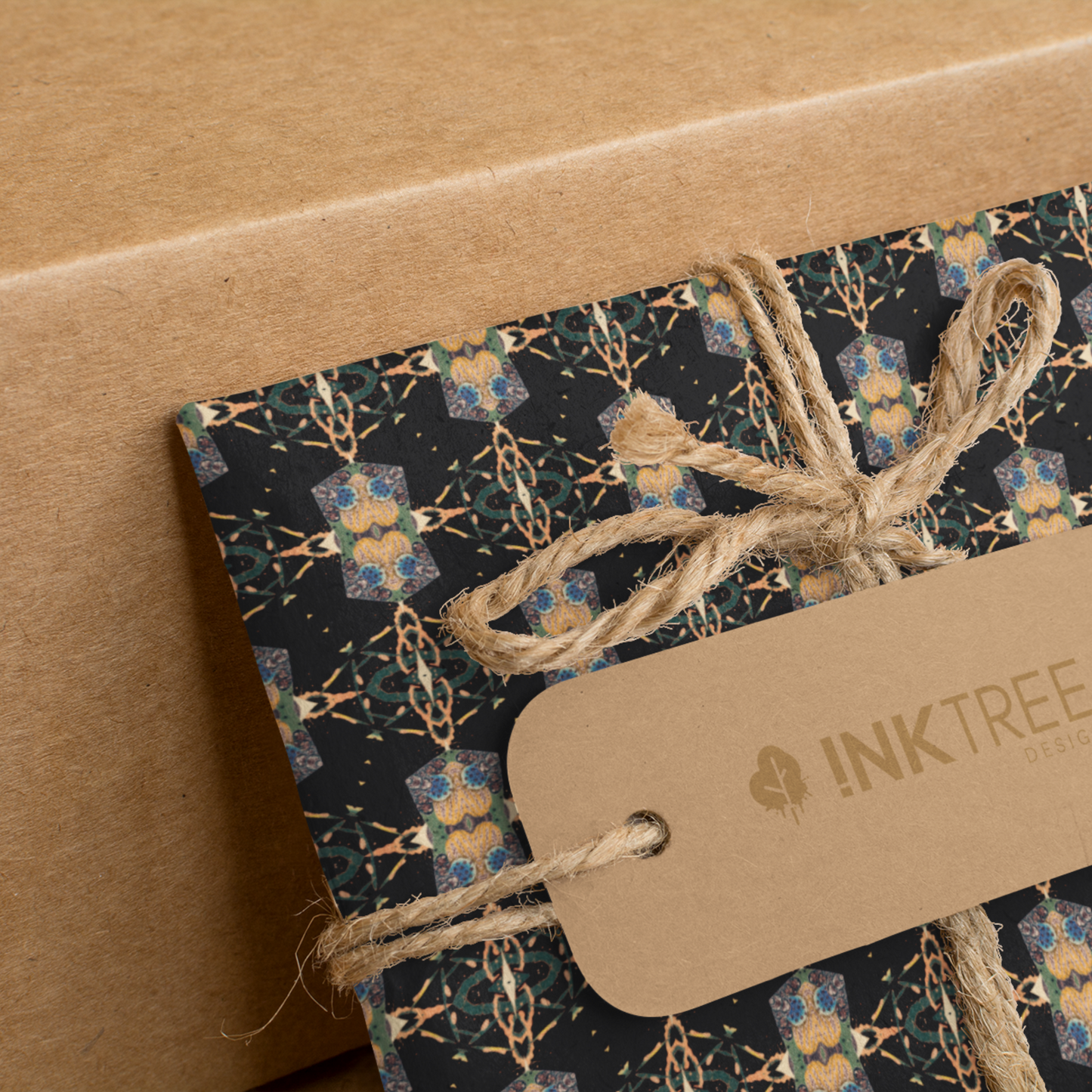 A present wrapped with a gold, black, blue, green and white oriental looking pattern, tied with brown string with a brown paper tag with ink tree design logo on it, leaning on a brown paper background.