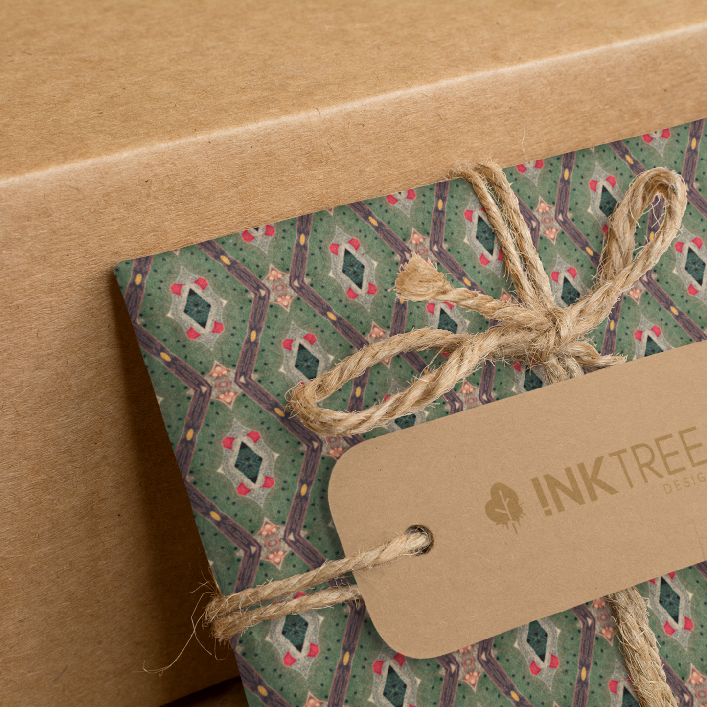 A present wrapped with a gold, black, green red and white oriental looking pattern, tied with brown string with a brown paper tag with ink tree design logo on it, leaning on a brown paper background.