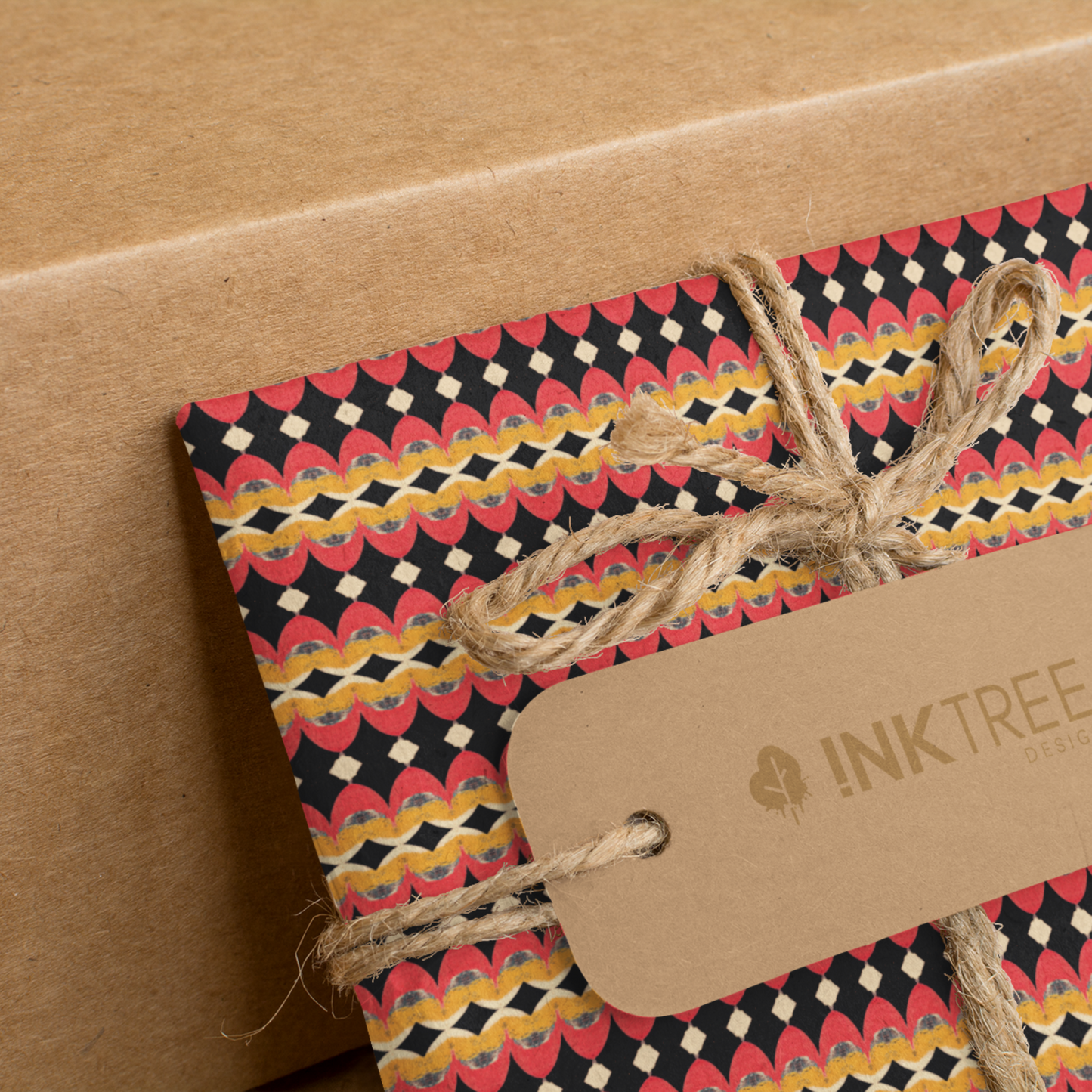 A present wrapped with an orange, black, white and yellow horizontal pattern, tied with brown string with a brown paper tag with ink tree design logo on it, leaning on a brown paper background.