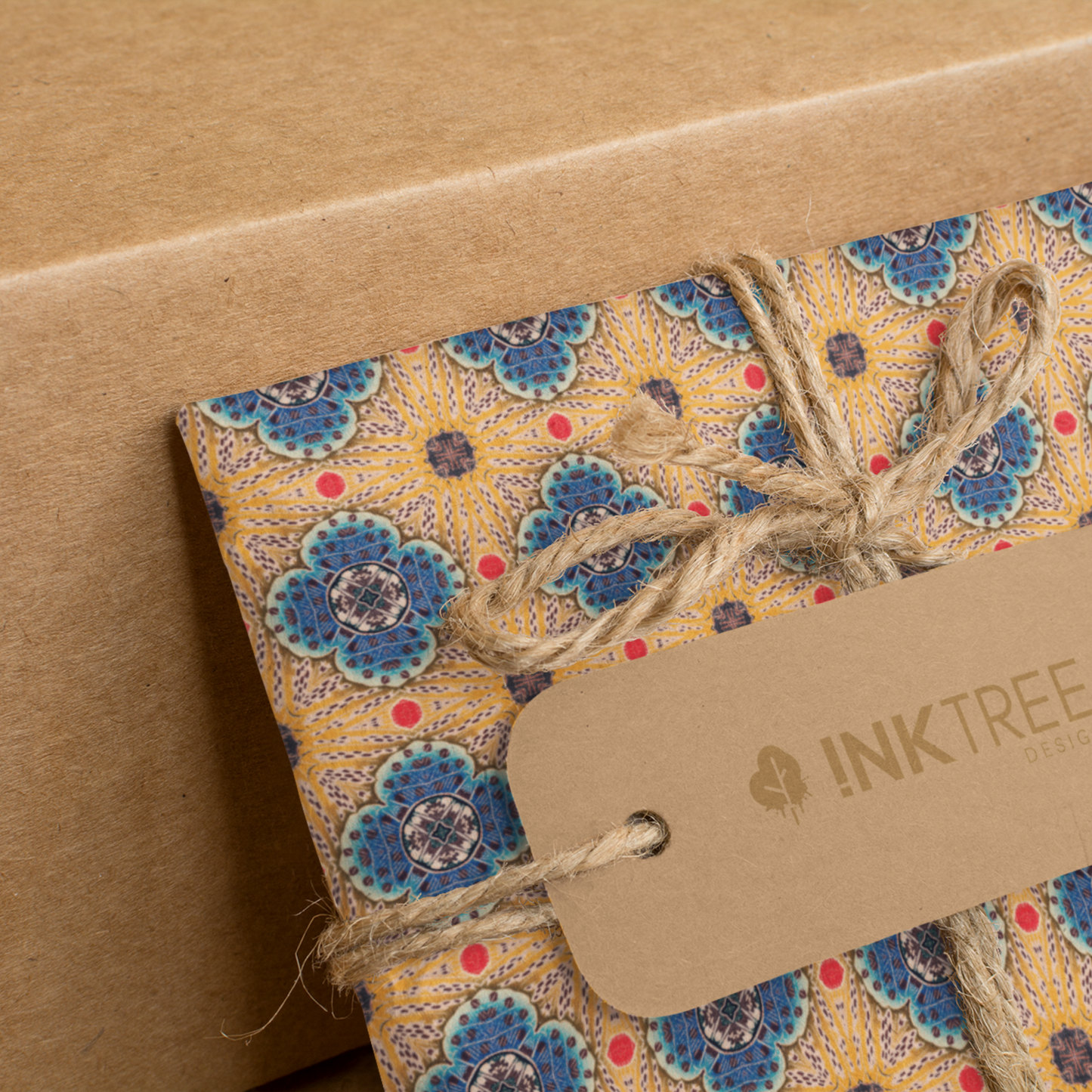 A present wrapped with a gold, black, blue, red and white oriental looking pattern, tied with brown string with a brown paper tag with ink tree design logo on it, leaning on a brown paper background.