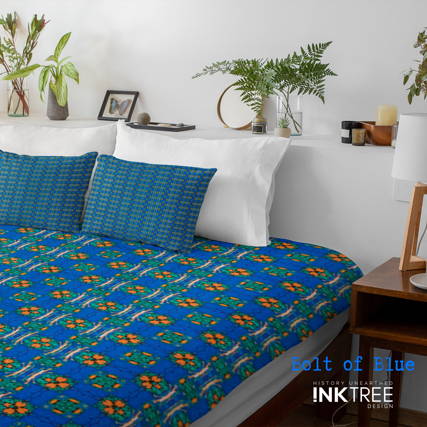 A doona cover and front pillows with an orange, white, black, blue and green floral pattern on a bed with a white wall, white pillows and white backboard background. There is a lamp with a white shade and wood base and plants and a small round metal mirror on the back board. There is also a bolt of blue, history unearthed ink tree design logo in the bottom right hand corner on it.