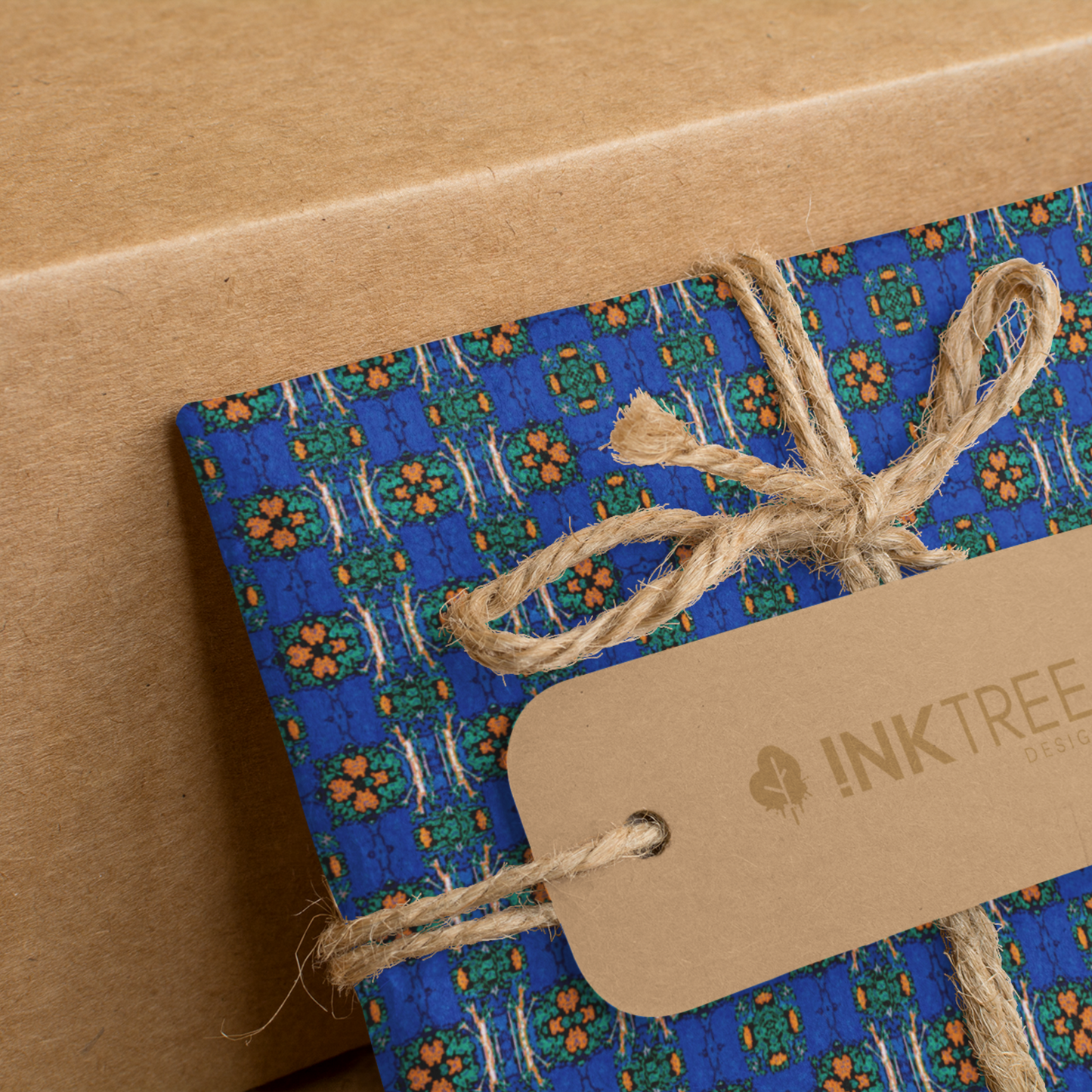 A present wrapped with an orange, white, black, blue and green floral pattern, tied with brown string with a brown paper tag with ink tree design logo on it, leaning on a brown paper background.