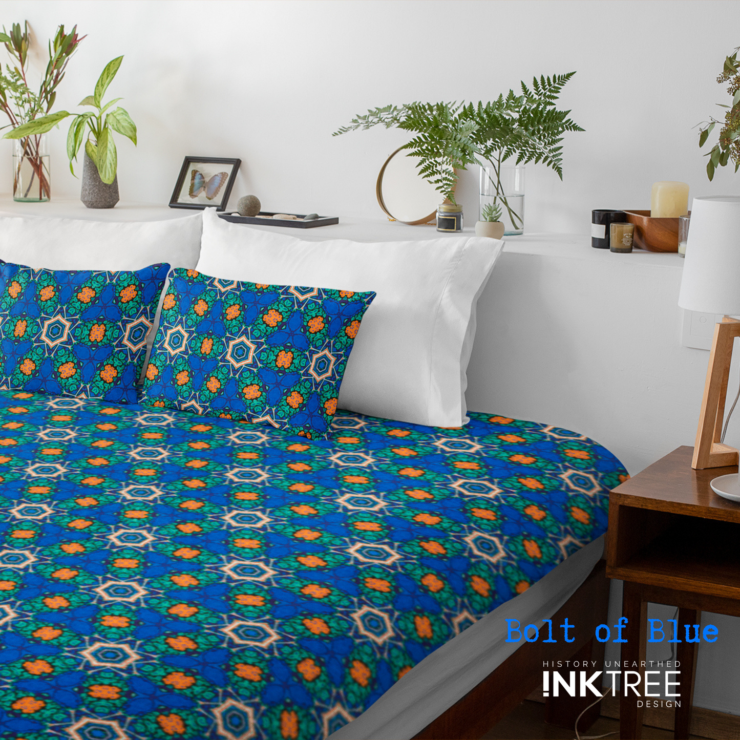 A doona cover and front pillows with an orange, white, black, blue and green pattern on a bed with a white wall, white pillows and white backboard background. There is a lamp with a white shade and wood base and plants and a small round metal mirror on the back board. There is also a bolt of blue, history unearthed ink tree design logo in the bottom right hand corner on it.