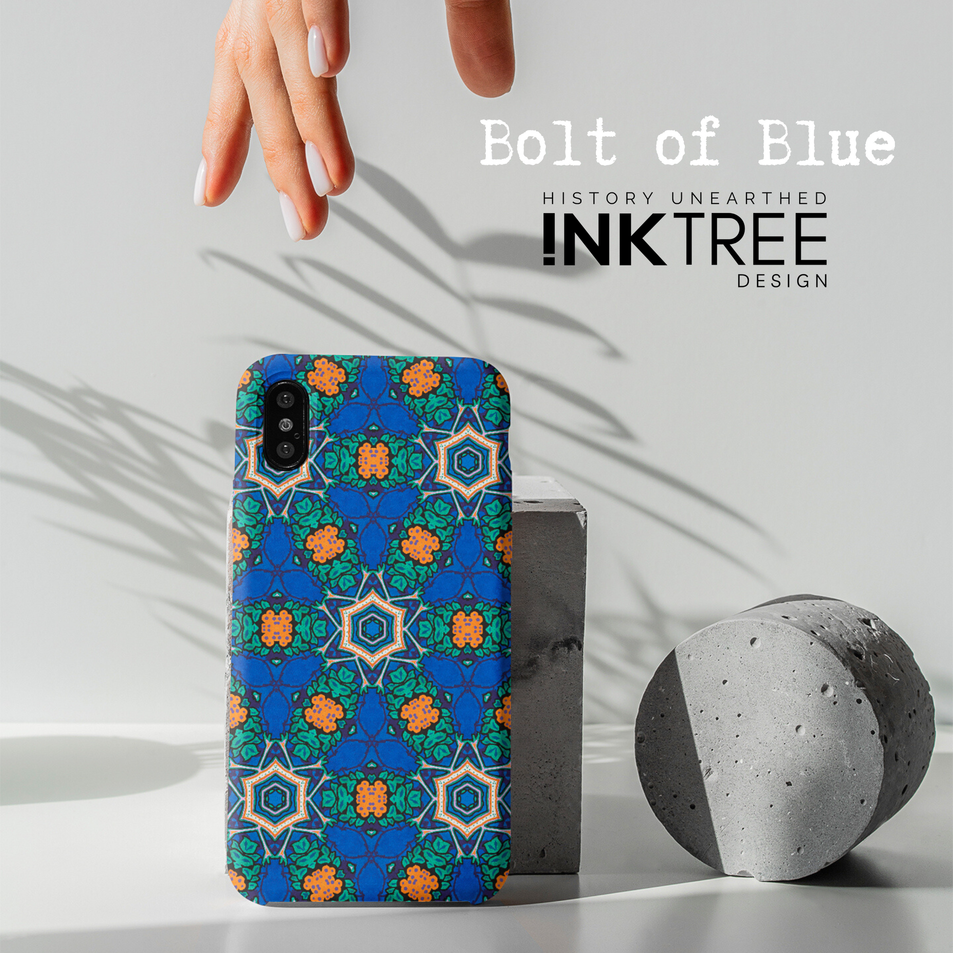 A female hand with white nail polish reaches down to a mobile phone with an orange, white, black, blue and green pattern the phone cover.  There is a shadow of a plant on a white wall background.  There is a grey concrete disk sitting on the table surface.