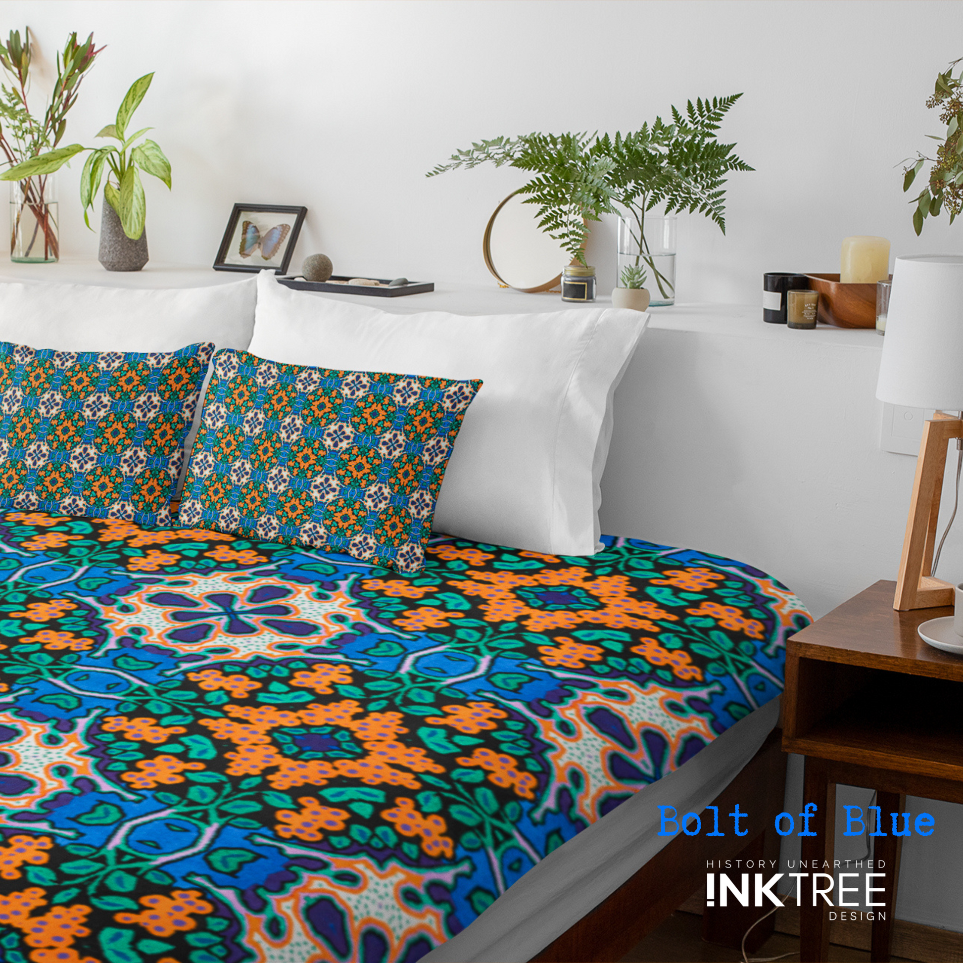 A doona cover and front pillows with an orange, white, black, blue and green pattern on a bed with a white wall, white pillows and white backboard background. There is a lamp with a white shade and wood base and plants and a small round metal mirror on the back board. There is also a bolt of blue, history unearthed ink tree design logo in the bottom right hand corner on it.