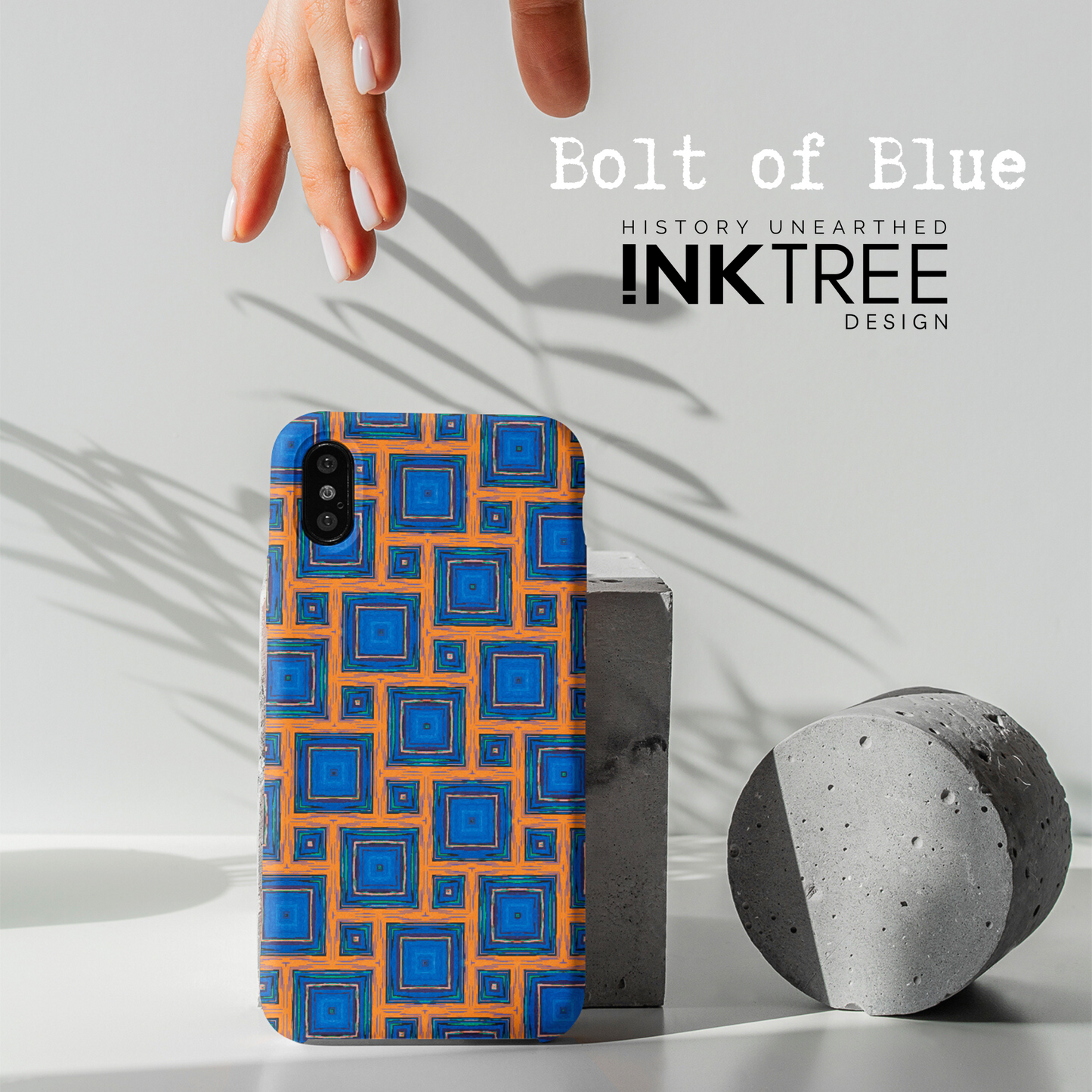 A female hand with white nail polish reaches down to a mobile phone with an orange, white, black, blue and green square pattern the phone cover.  There is a shadow of a plant on a white wall background.  There is a grey concrete disk sitting on the table surface.