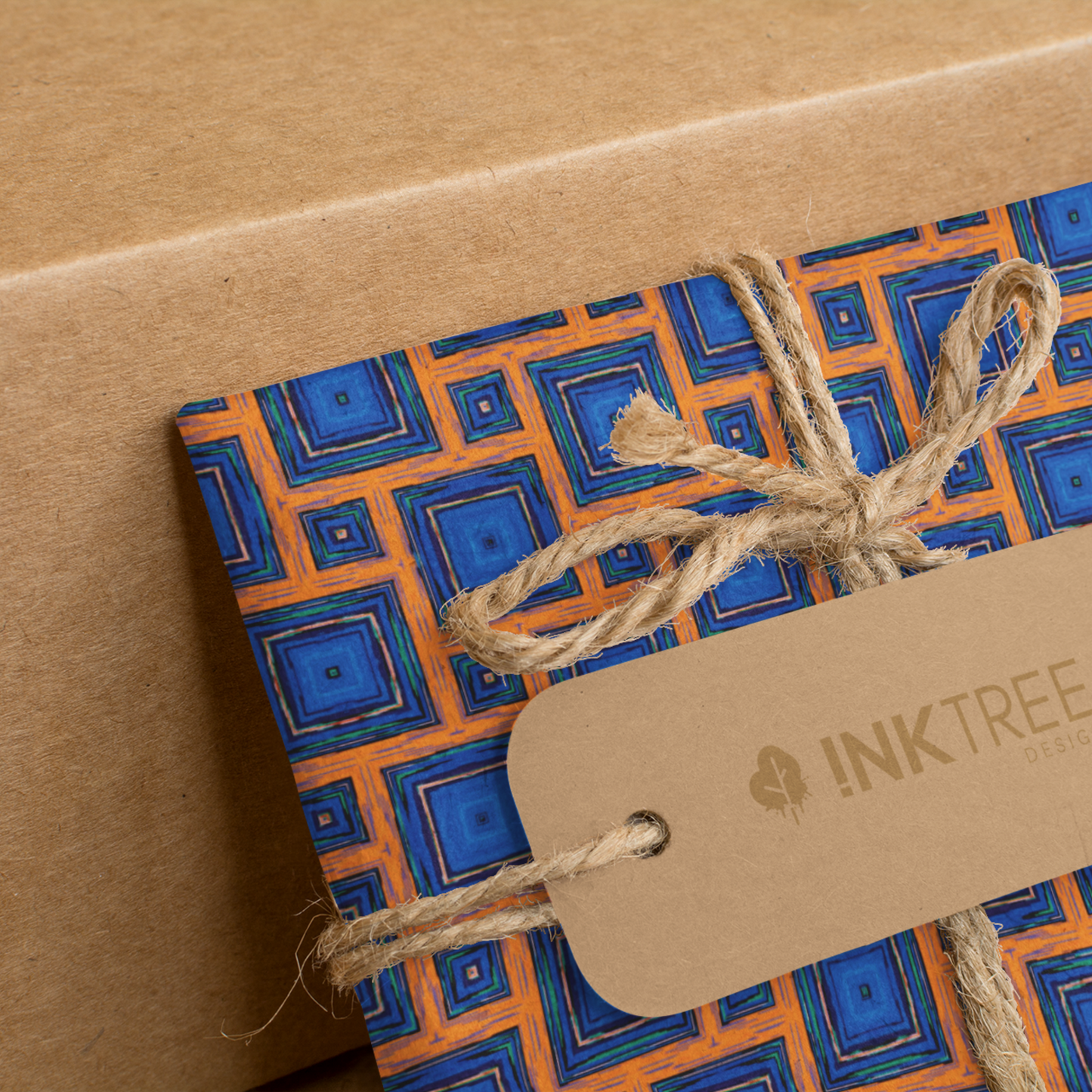 A present wrapped with an orange, white, black, blue and green square pattern, tied with brown string with a brown paper tag with ink tree design logo on it, leaning on a brown paper background.