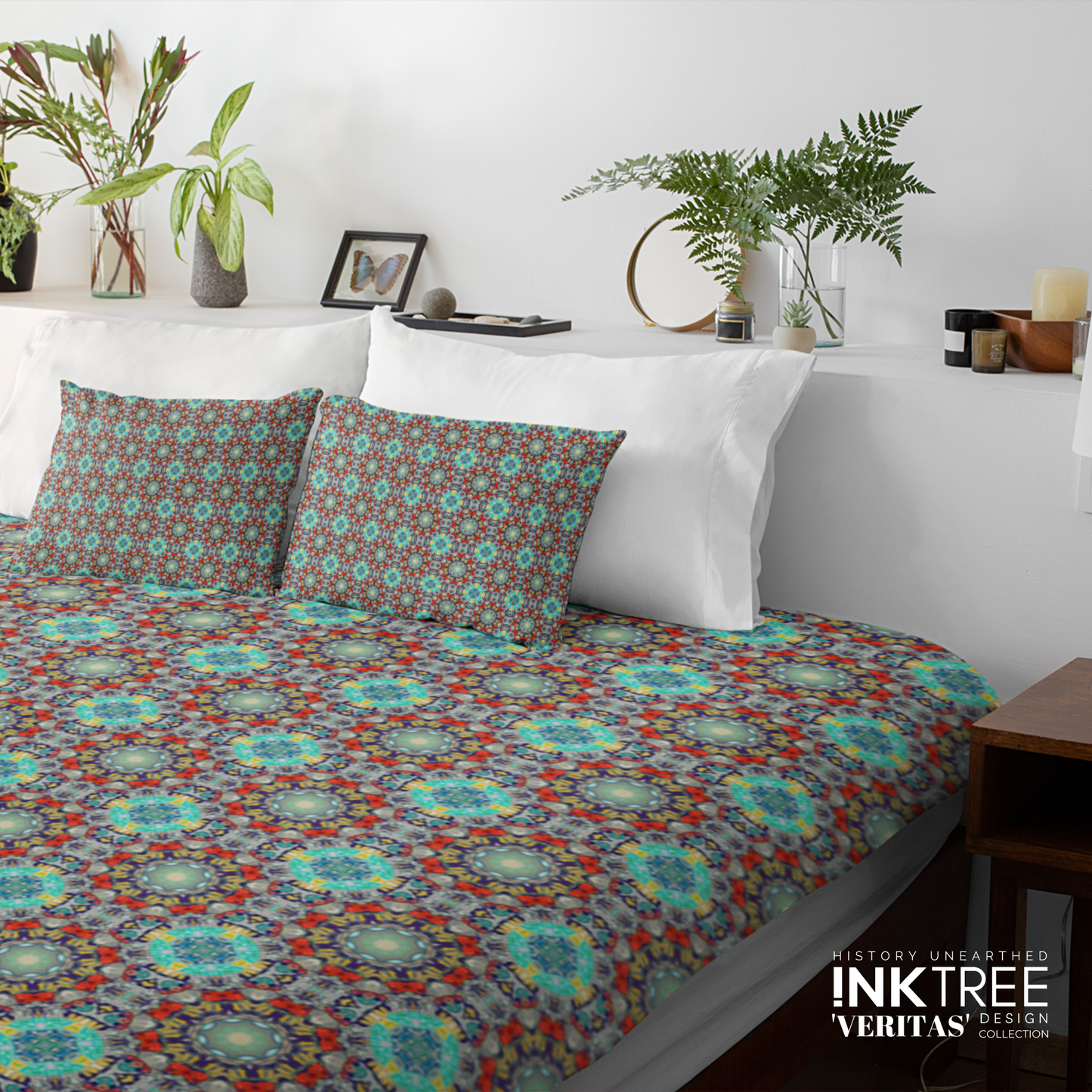 A doona cover and pillows with blue and aqua and red pattern against a white wall, leaning against white pillows and green plants in vases.