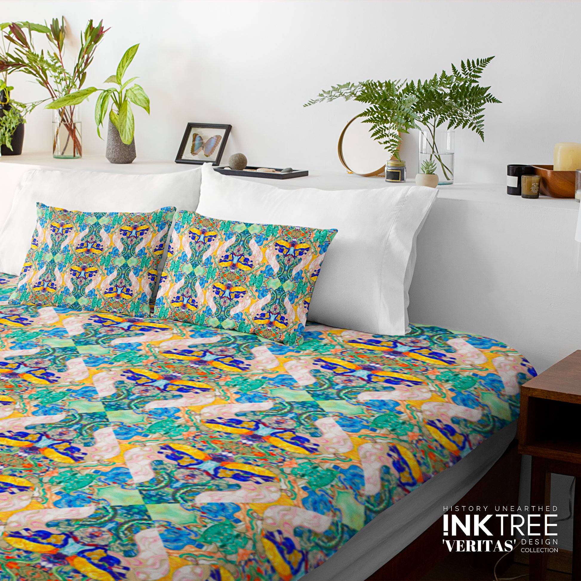 A doona cover and pillows with blue, green, gold, pattern on a white wall, leaning against white pillows and green plants in vases.
