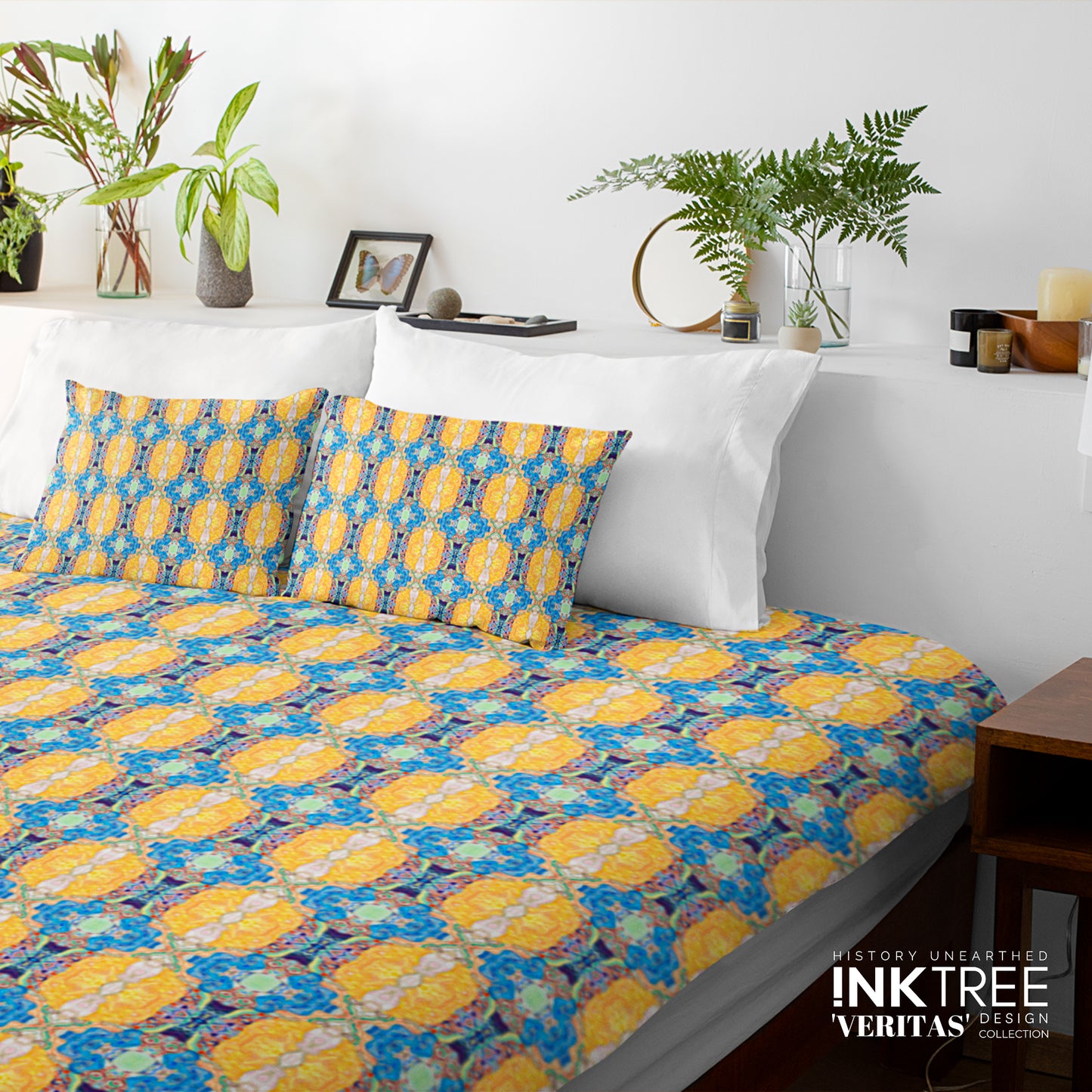 A doona cover and pillows with blue and yellow pattern against a white wall, leaning against white pillows and green plants in vases.