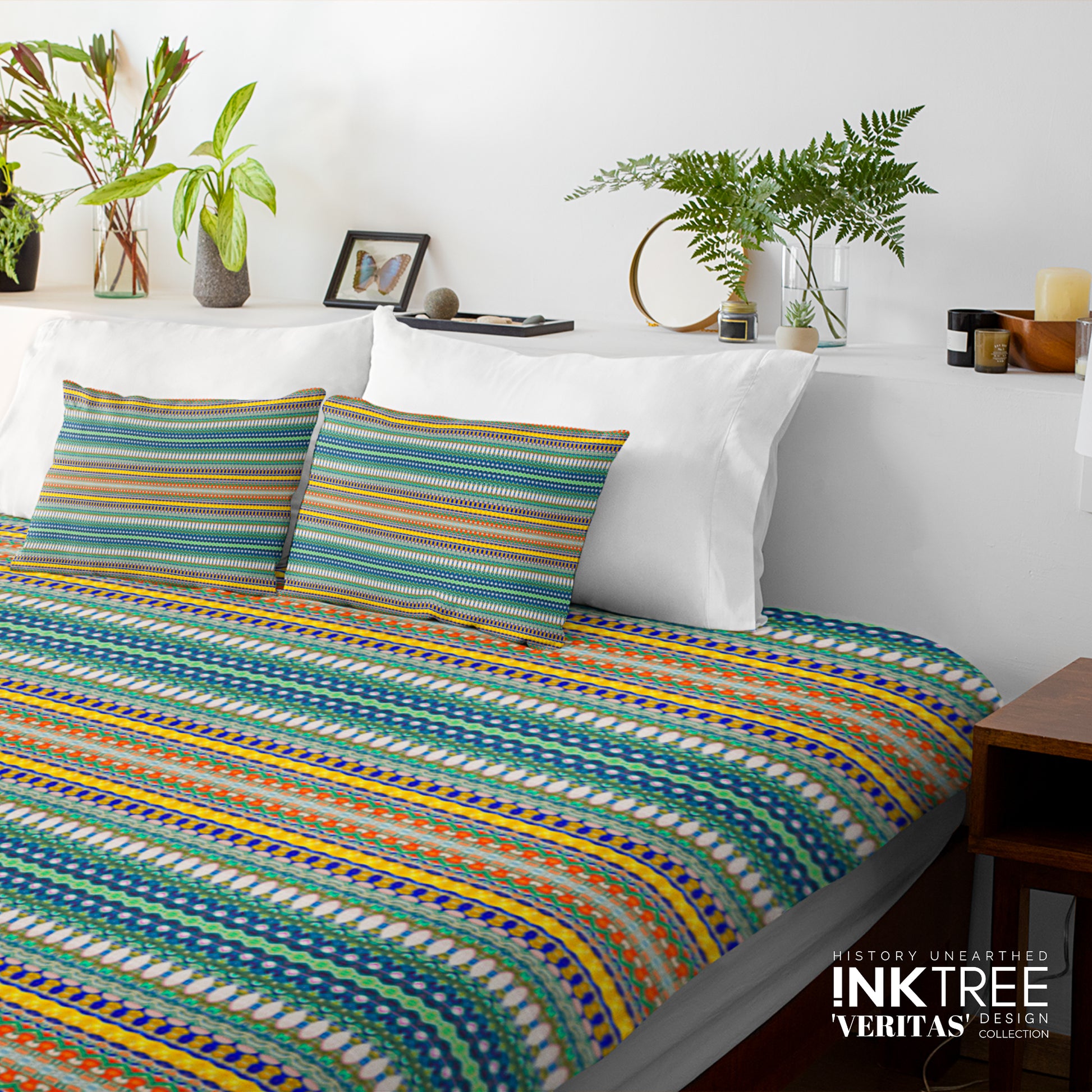 A doona cover and pillows with blue and yellow, green and orange pattern against a white wall, leaning against white pillows and green plants in vases.