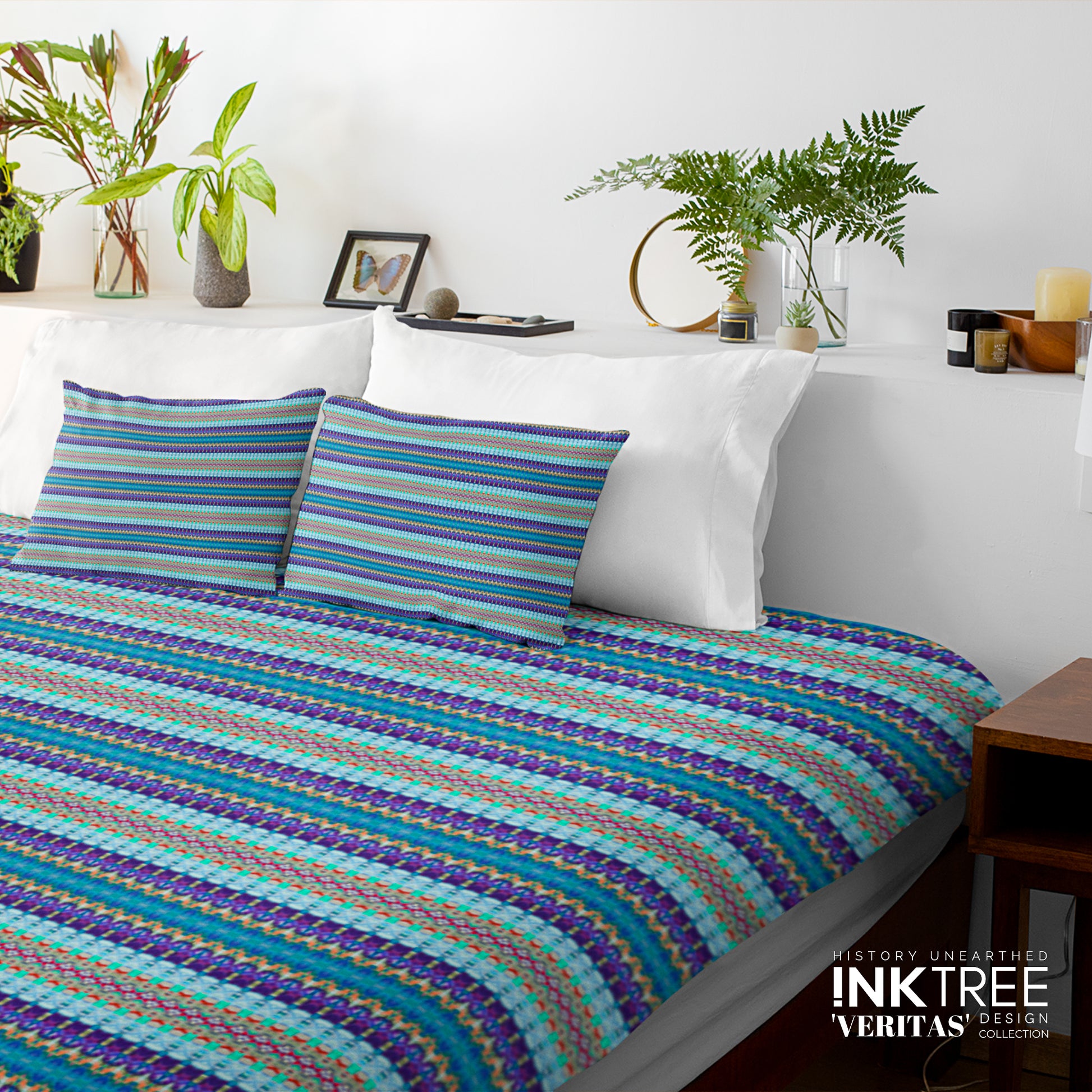 A doona cover and pillows with blue and green horizontal pattern against a white wall, leaning against white pillows and green plants in vases.