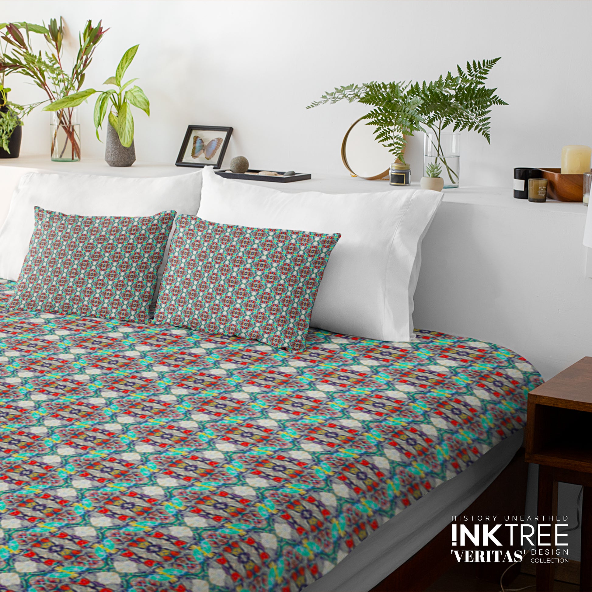 A doona cover and pillows with blue,red and green pattern against a white wall, leaning against white pillows and green plants in vases.
