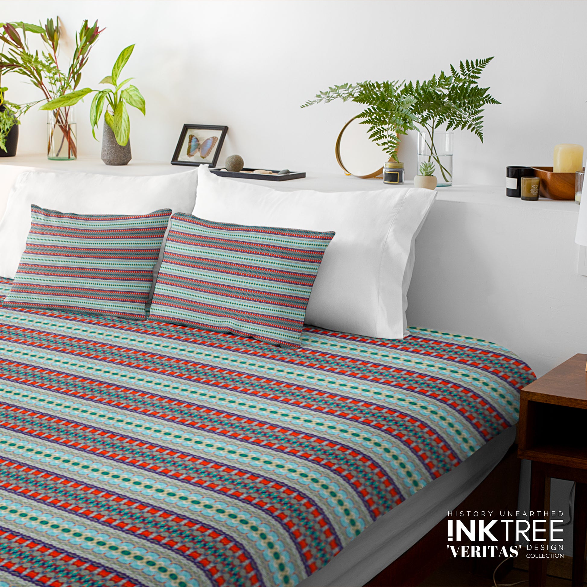 A doona cover and pillows with blue, red and green horizontal line pattern against a white wall, leaning against white pillows and green plants in vases.