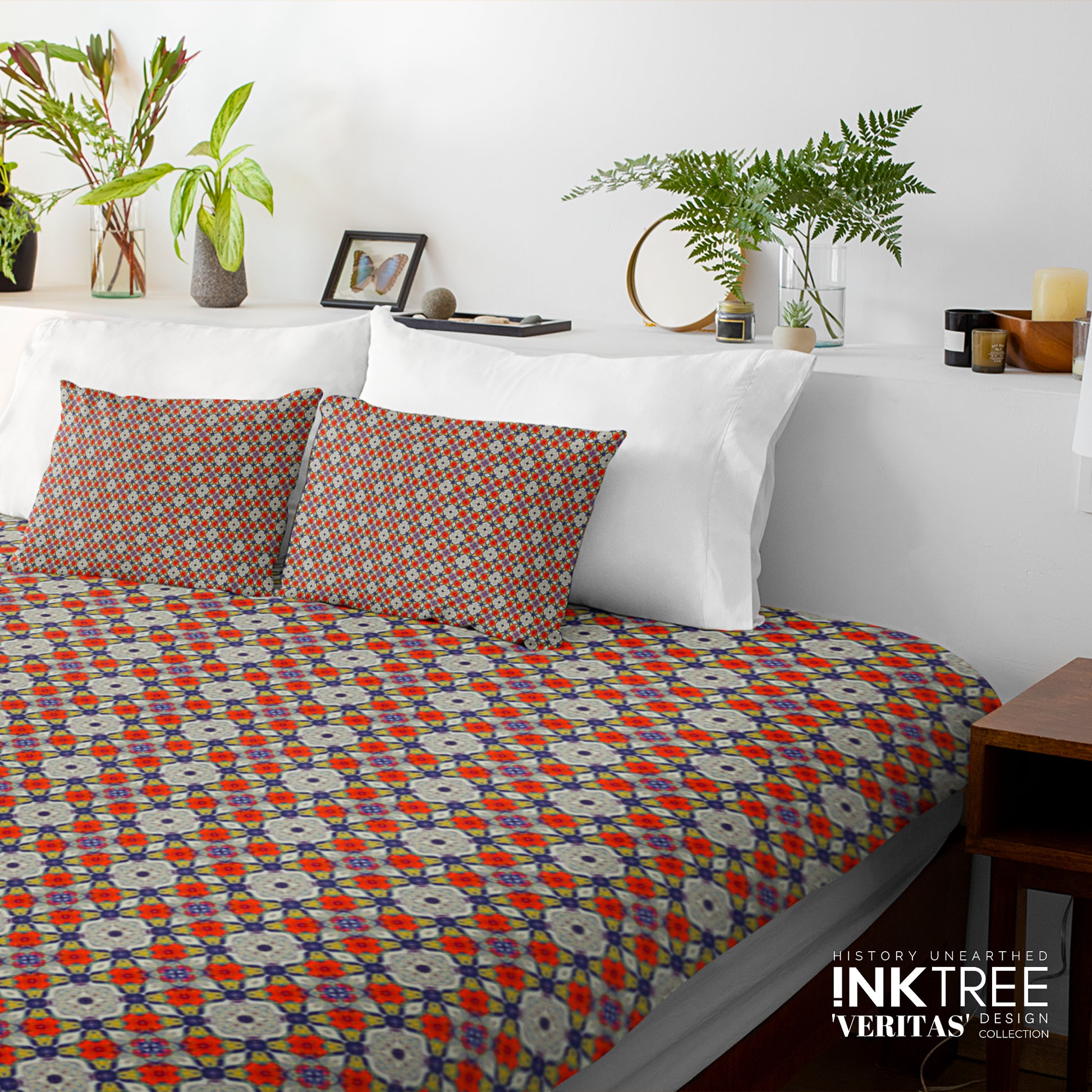 A doona cover and pillows with blue, grey and red floral pattern against a white wall, leaning against white pillows and green plants in vases.