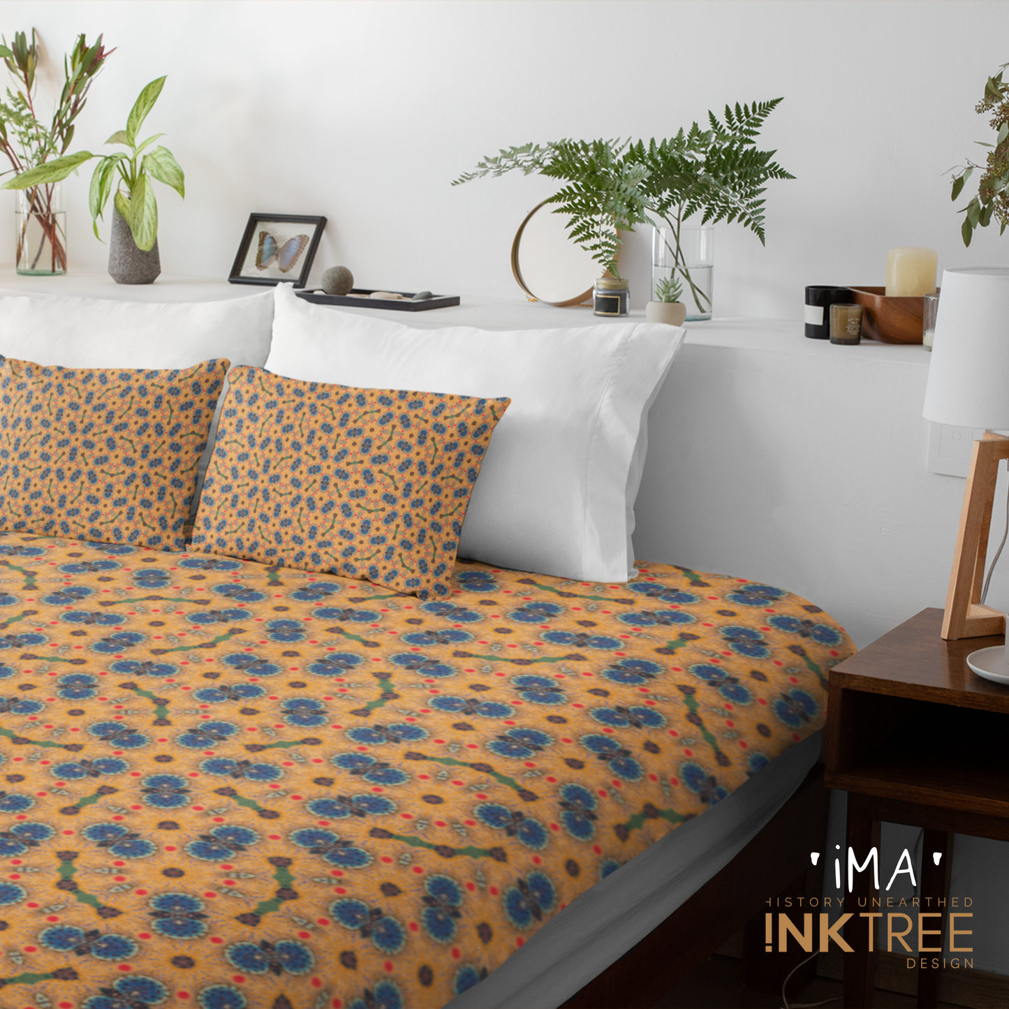 A quilt or doona cover and front pillows with a gold, black, blue, red, green and white oriental looking pattern on a bed with a white wall, white pillows and white backboard background. There is a lamp with a white shade and wood base and plants and a small round metal mirror on the back board. There is also a ima, history unearthed ink tree design logo in the bottom right hand corner on it.