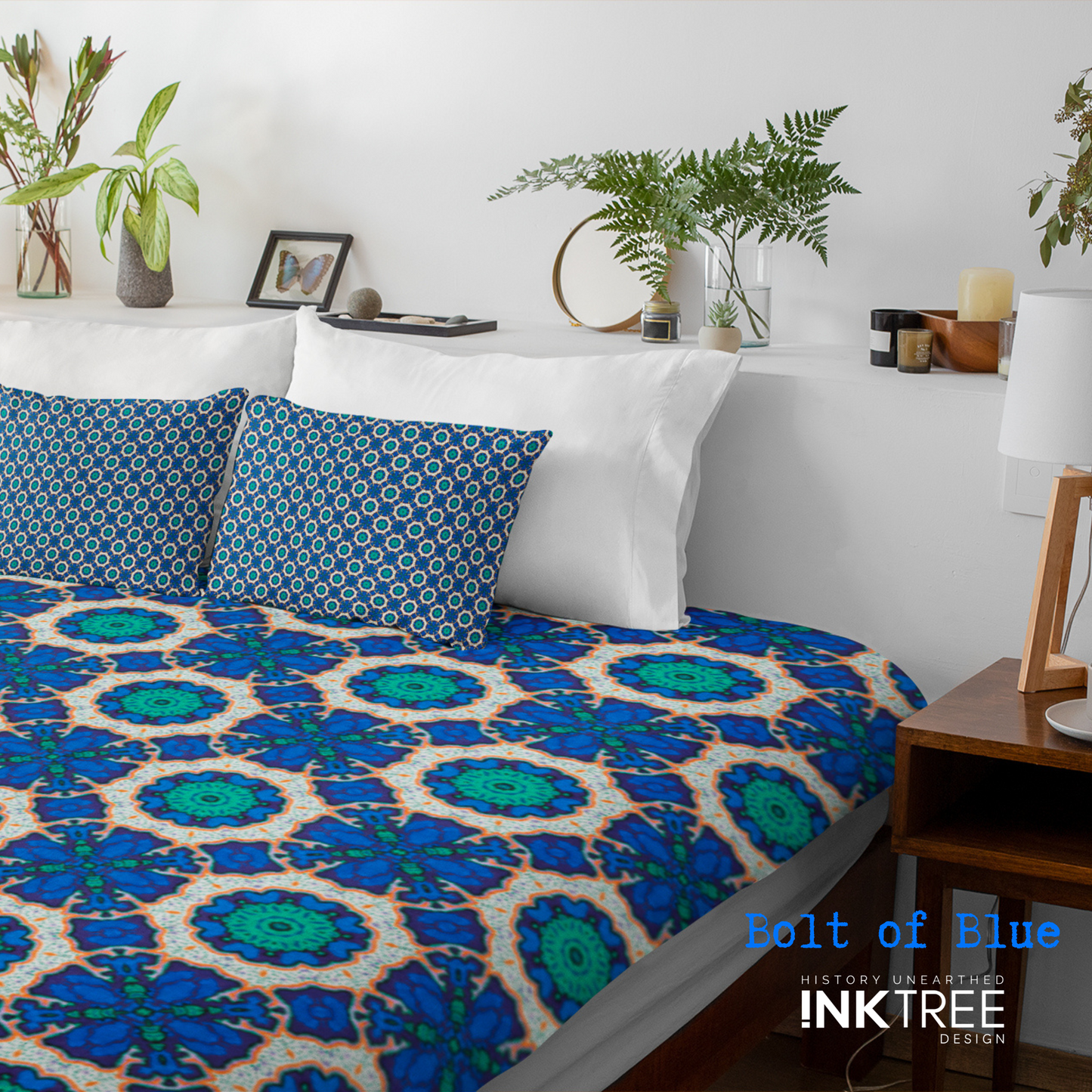 A doona cover and front pillows with an orange, white, black, blue and green floral pattern on a bed with a white wall, white pillows and white backboard background. There is a lamp with a white shade and wood base and plants and a small round metal mirror on the back board. There is also a bolt of blue, history unearthed ink tree design logo in the bottom right hand corner on it.