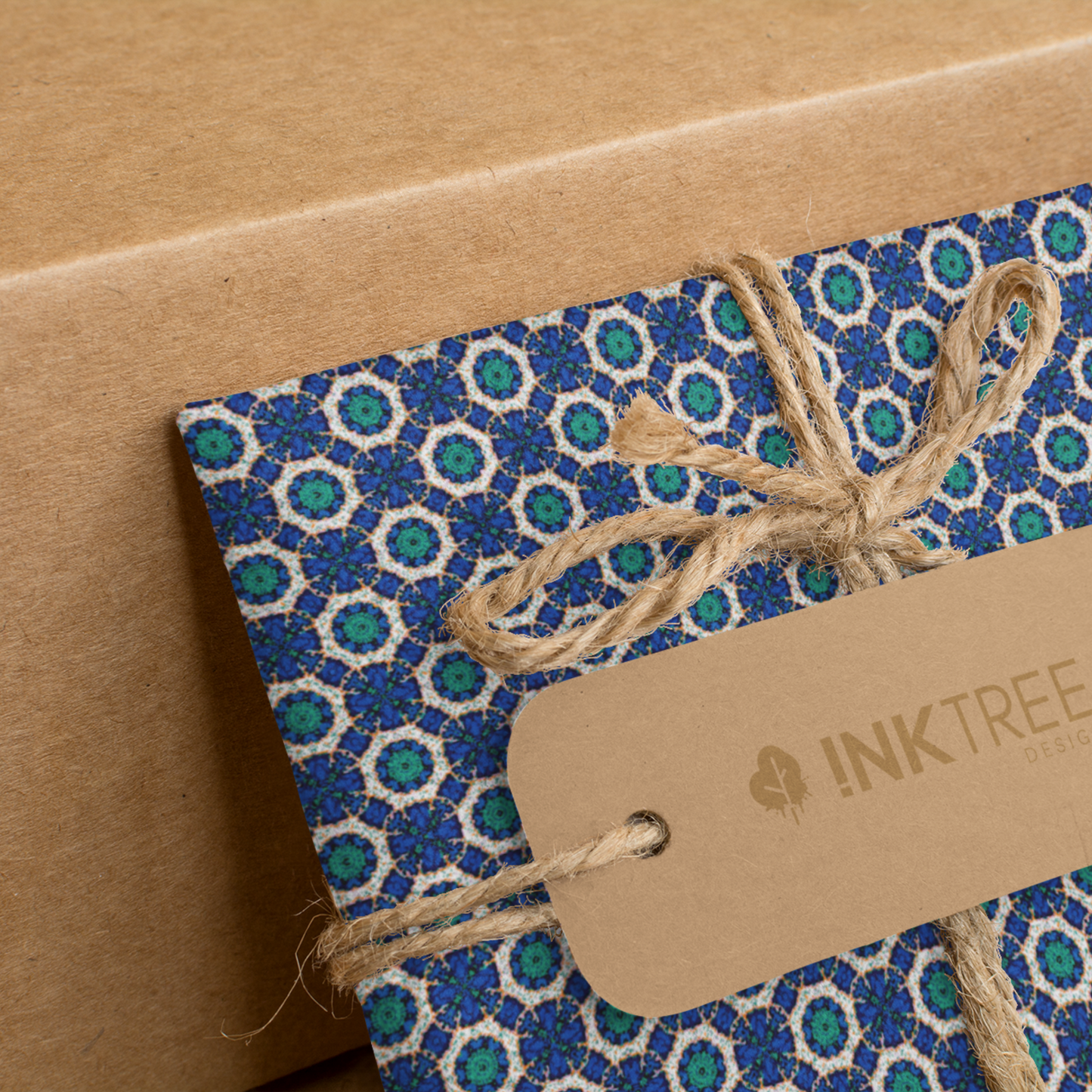 A present wrapped with an orange, white, black, blue and green floral pattern, tied with brown string with a brown paper tag with ink tree design logo on it, leaning on a brown paper background.