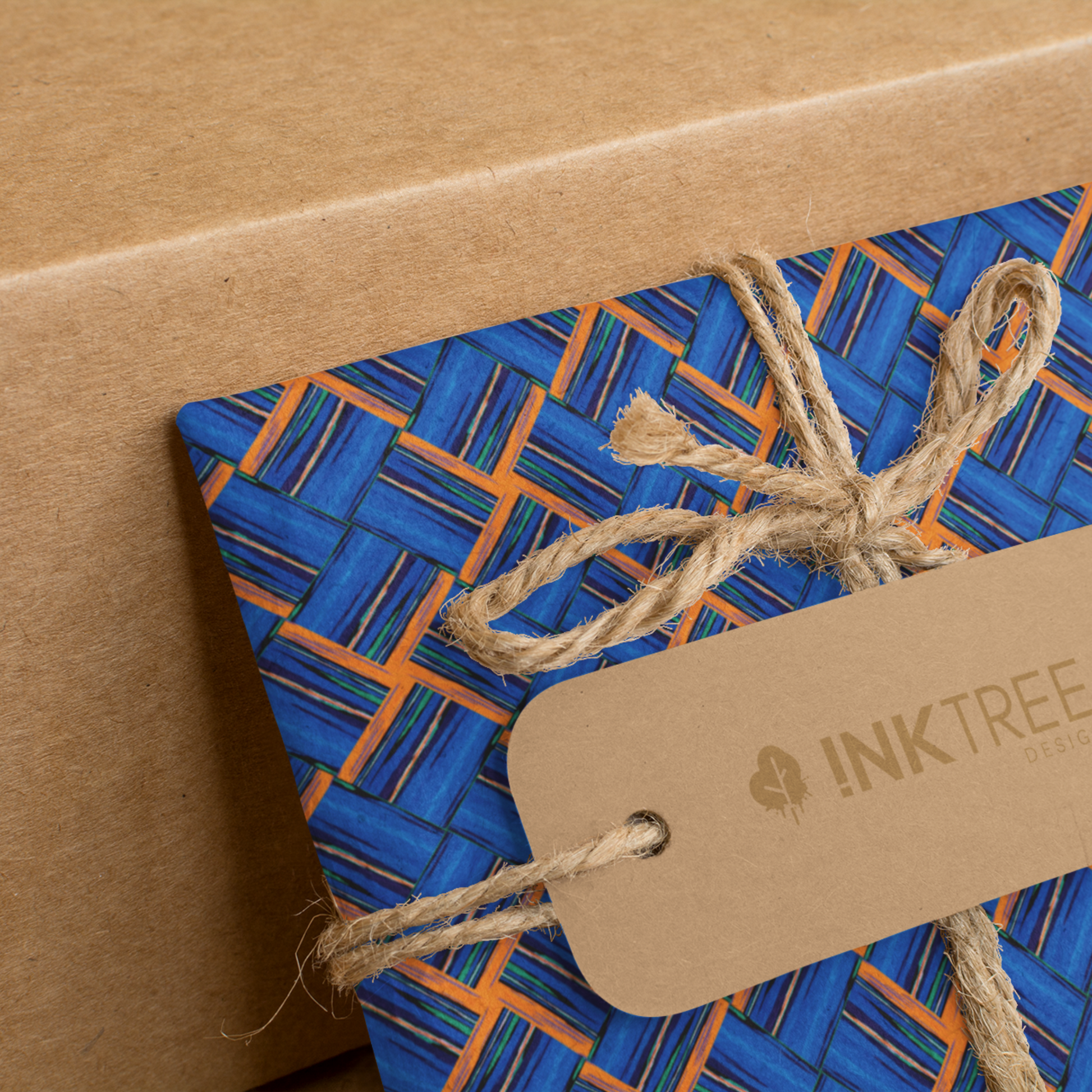 A present wrapped with an orange, white, black, blue and green square pattern, tied with brown string with a brown paper tag with ink tree design logo on it, leaning on a brown paper background.