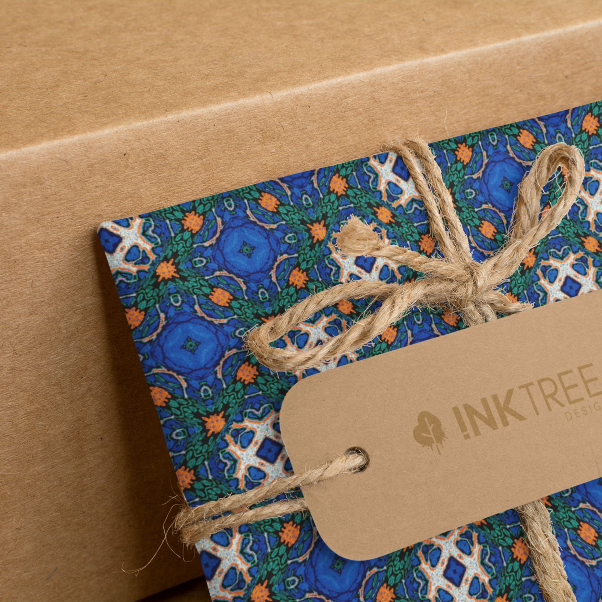A present wrapped with an orange, white, black, blue and green pattern, tied with brown string with a brown paper tag with ink tree design logo on it, leaning on a brown paper background.