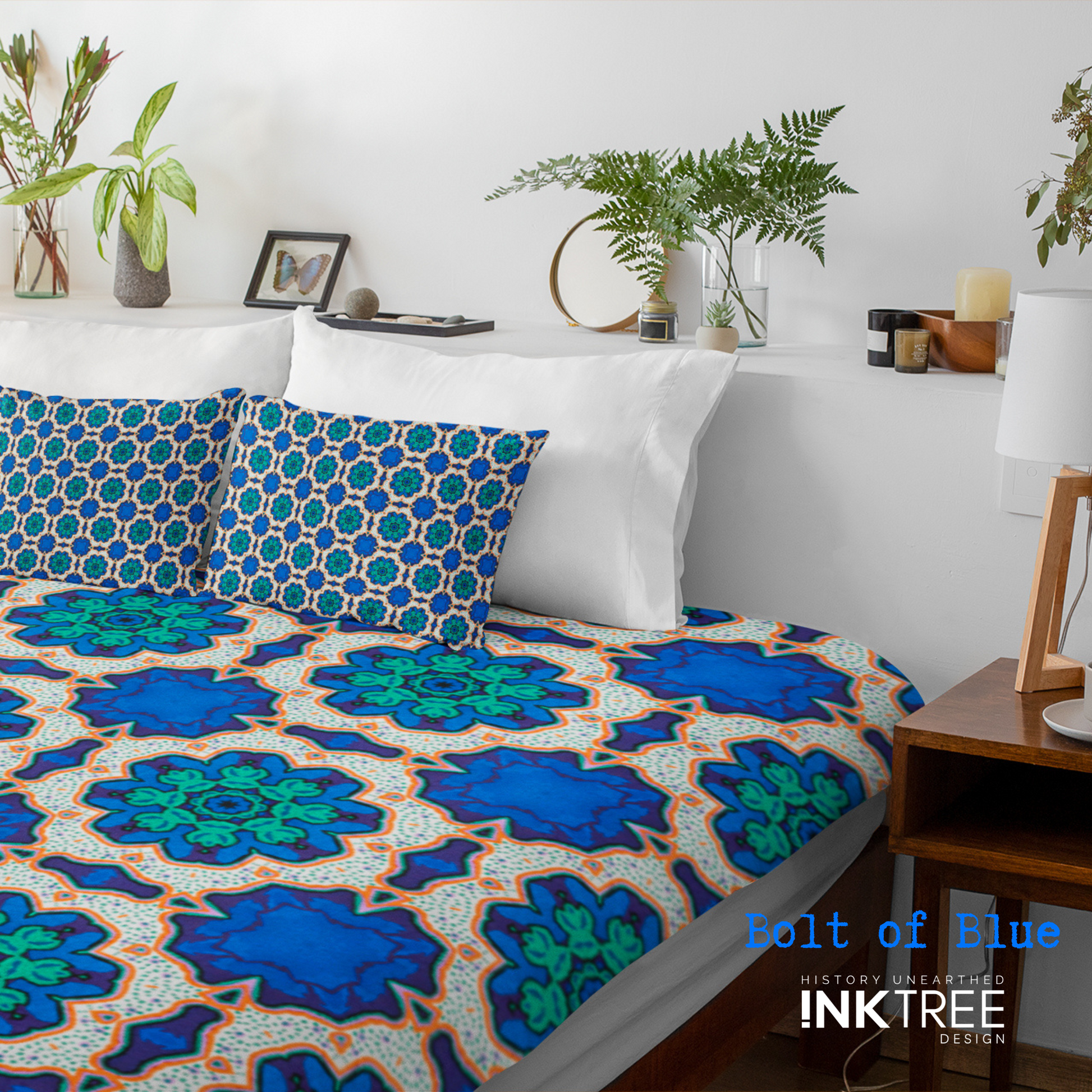 A doona cover and front pillows with an orange, white, black, blue and green floral pattern on a bed with a white wall, white pillows and white backboard background. There is a lamp with a white shade and wood base and plants and a small round metal mirror on the back board. There is also a bolt of blue, history unearthed ink tree design logo in the bottom right hand corner on it.