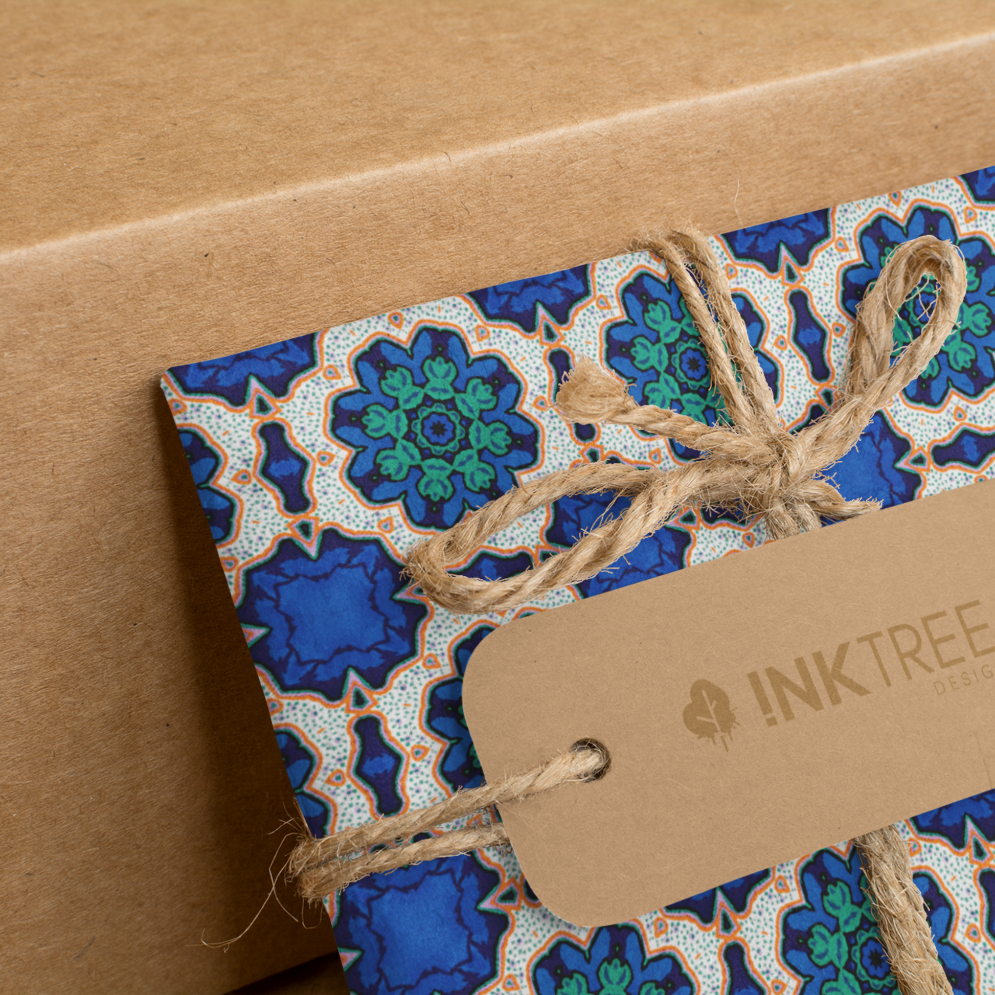  A present wrapped with an orange, white, black, blue and green floral pattern, tied with brown string with a brown paper tag with ink tree design logo on it, leaning on a brown paper background.